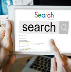 A finger touches a tablet screen displaying a Google search bar with the word "Search."