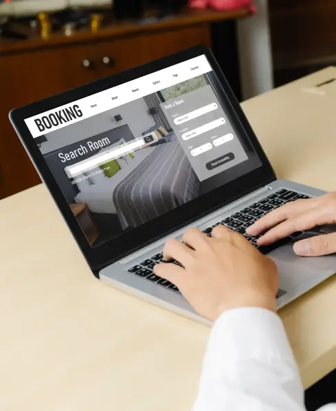 A person types on a laptop displaying a hotel website with a prominent "BOOKING" and "Search Room" interface